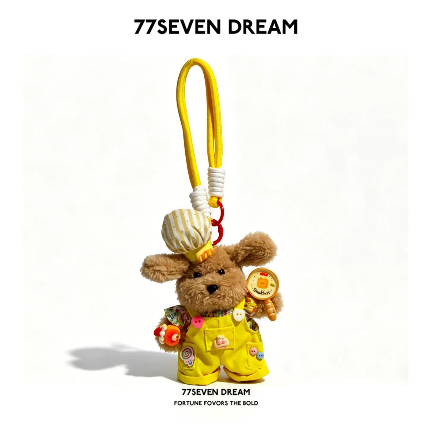 Brown plush dog charm in yellow overalls with colorful embellishments and yellow hanging cord