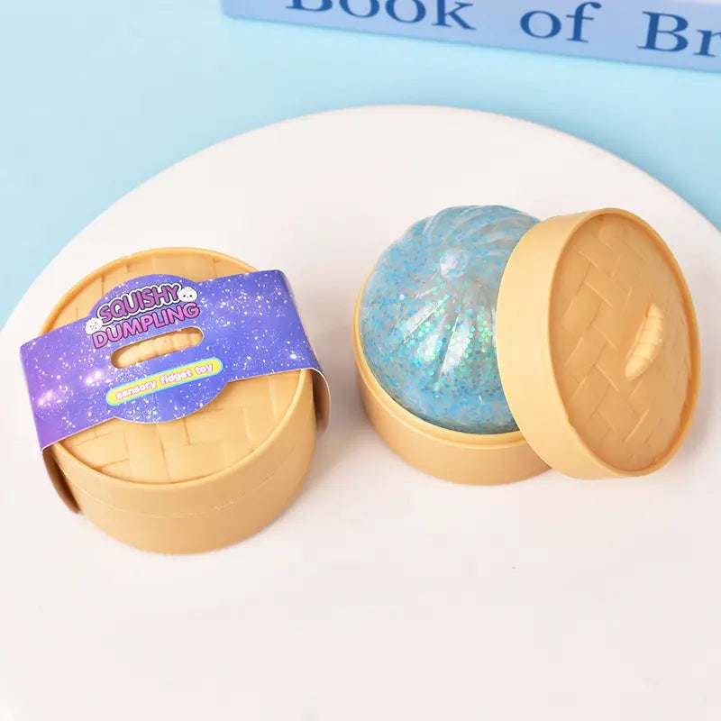 The ZoDoll Glitter Squishy Dumpling Sensory Fidget Toy with Steamer, a blue glittery dumpling-shaped toy, is shown on a white surface alongside its beige steamer container.