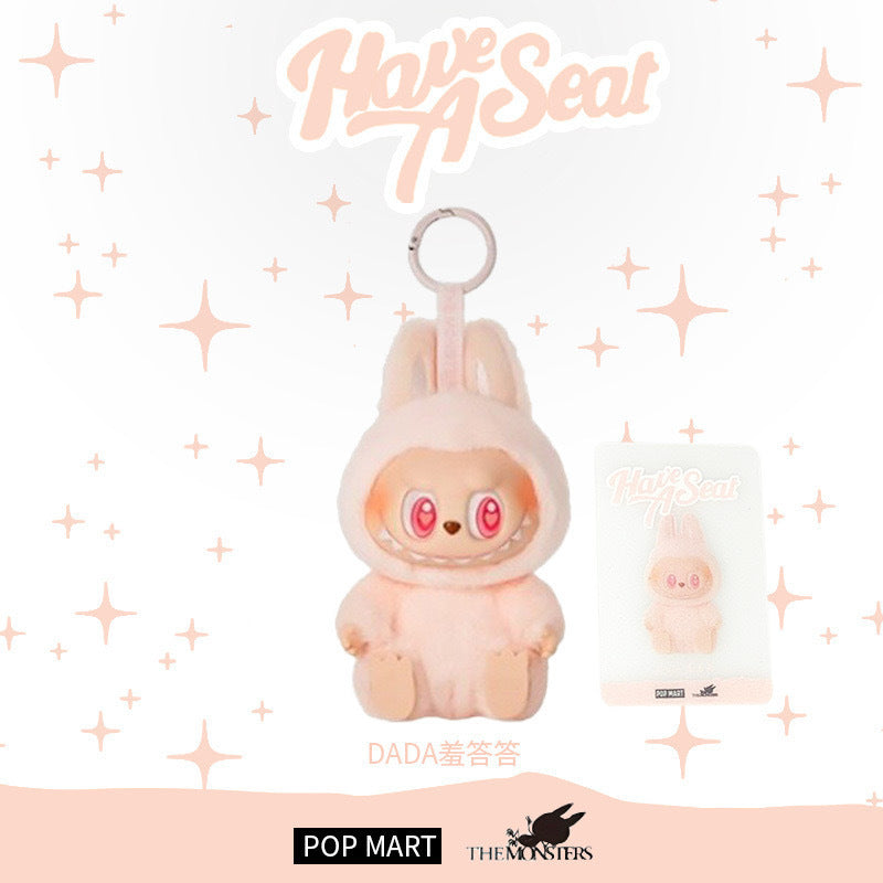 The Labubu "Dada" Plush Keychain – Have a Seat Series Pink Heart-Eye Bunny by POP MART features a kawaii pastel pink bunny with big eyes, shown in packaging with "Have A Seat" text—a perfect collectible for Pop Mart fans.