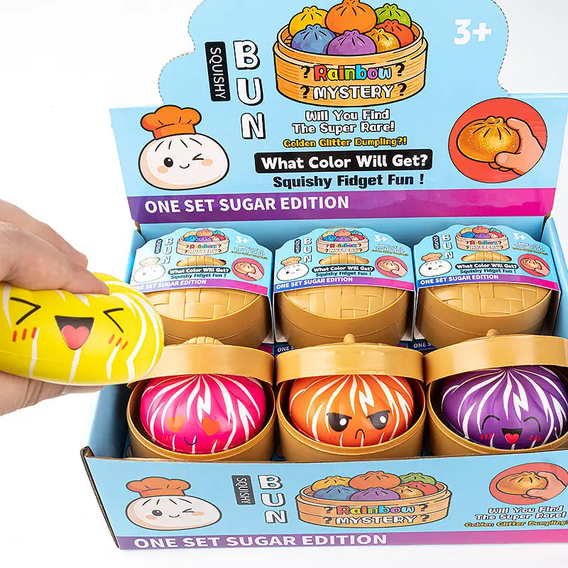 A hand squeezes ZoDoll’s Rainbow Mystery Squishy Bun Fidget Toy with Steamer – Sugar Edition near an open box showing colorful bun toys in gold containers, set against a vibrant gradient background.