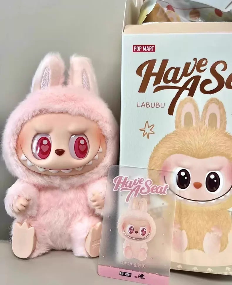 The Labubu “Dada” Plush Keychain – Have a Seat Series Pink Heart-Eye Bunny by POP MART sits beside its matching box and collectible card, both featuring the same Pop Mart The Monsters character.