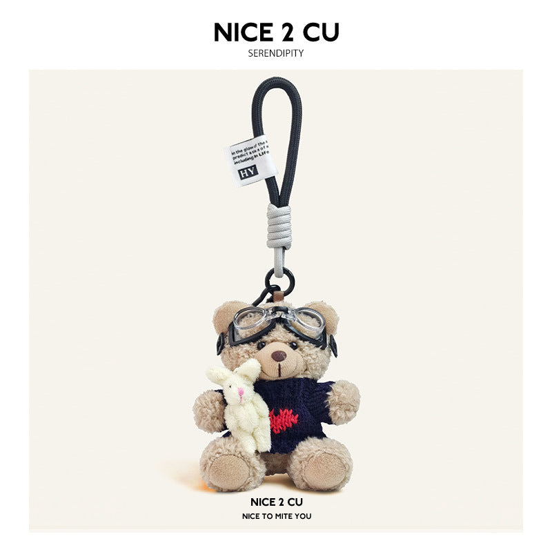 Beige plush teddy bear keychain in dark navy knit sweater with small heart motif and aviator goggles, holding a white rabbit