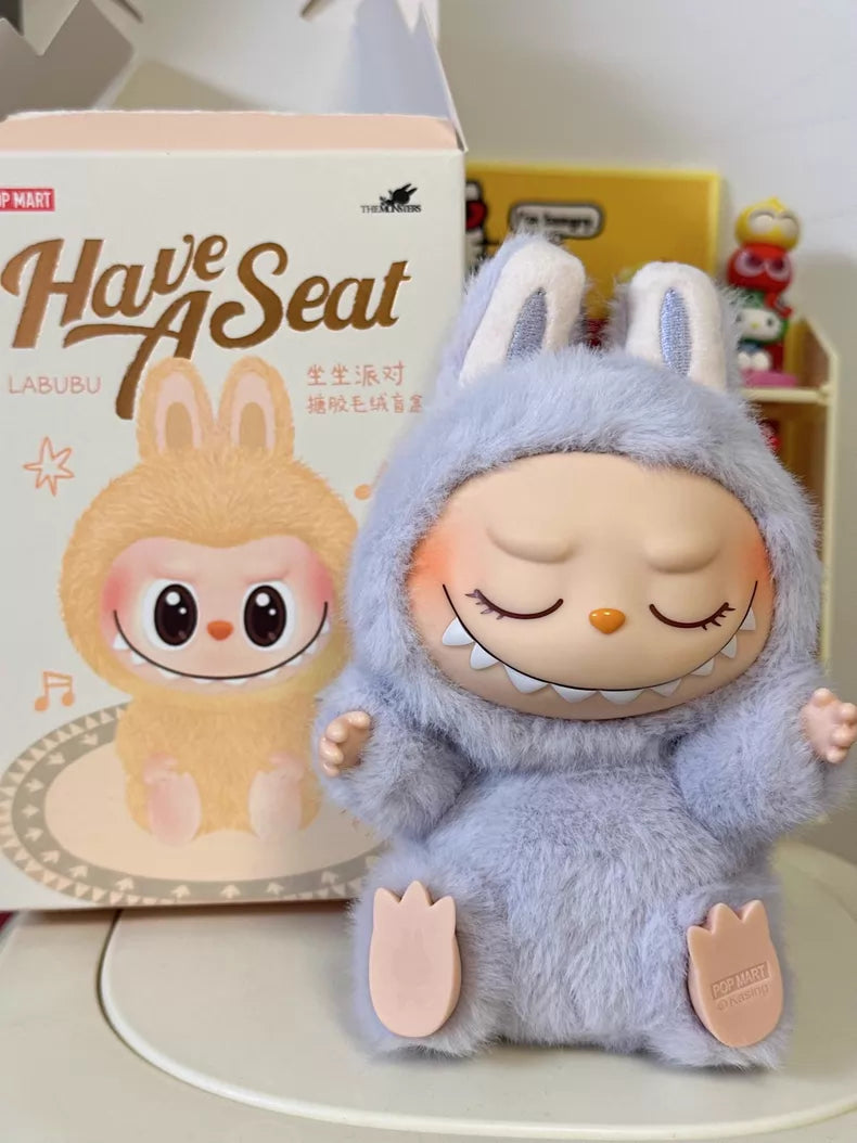 A POP MART Labubu "Zizi" Plush Keychain from The Monsters Have a Seat Series sits with closed eyes and a wide grin, displayed before its illustrated packaging.