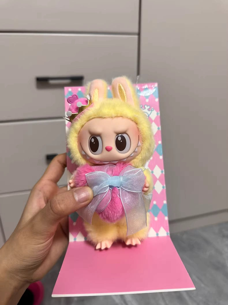 A hand holds the POP MART Labubu Let's Checkmate Series – Pink Heart Bunny Plush Vinyl Doll, dressed in a yellow bunny suit and holding a pink heart with a blue ribbon, in front of light drawers—a whimsical collectible for any space.