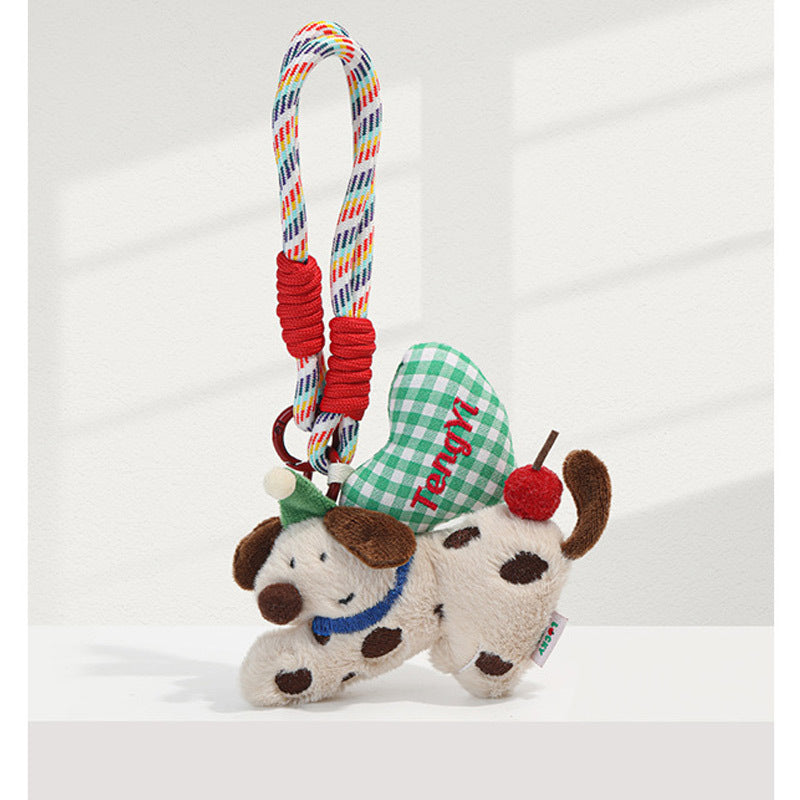 Cream plush dog charm keychain with brown spots, green party hat, and green gingham heart on braided multicolor cord