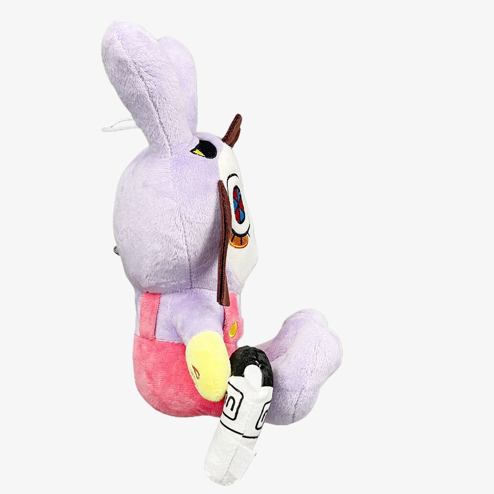 A side view of The Amazing Digital Circus Inspired Plush Toy—a 9.8-inch bunny ear cartoon doll with purple fur, big eyes, pink overalls, a white mask with blue and red details, holding a white object with black markings.