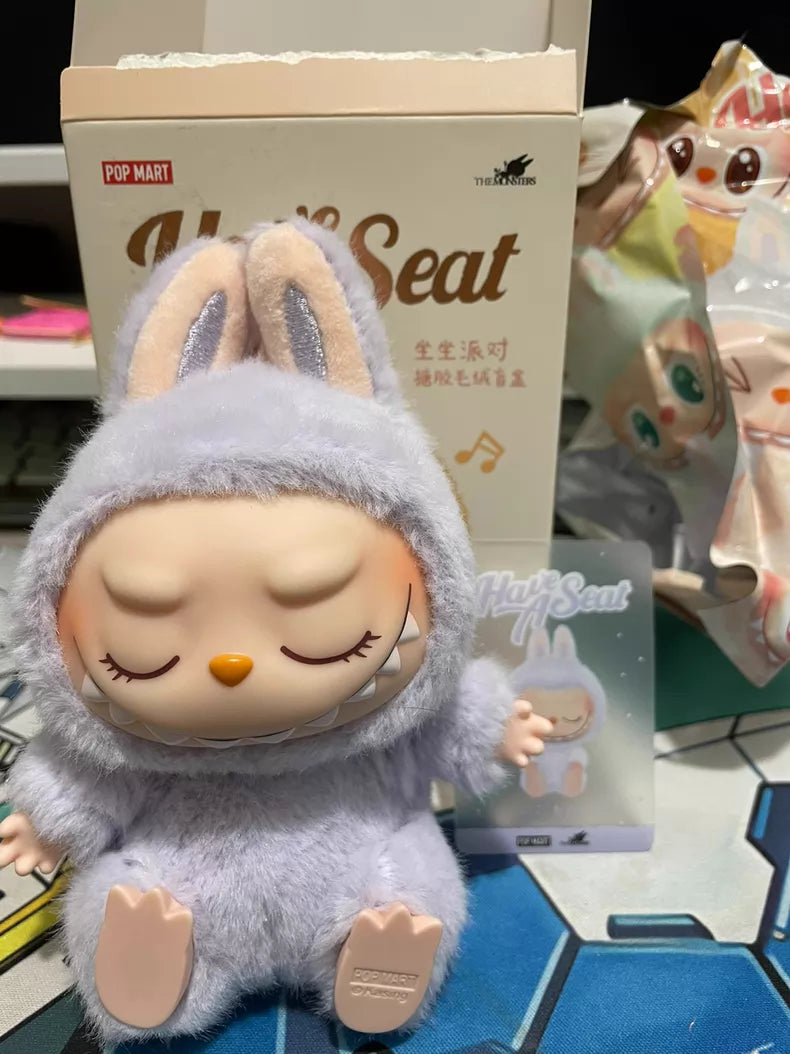 The Labubu “Zizi” Plush Keychain from the POP MART Have a Seat Series is shown with closed eyes and bunny ears, sitting in front of branded packaging and a card from The Monsters Collection.