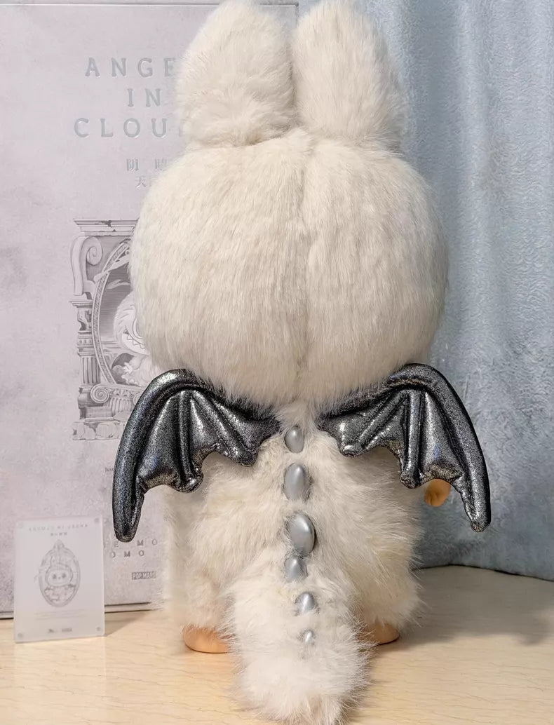 The POP MART Labubu Angel in Clouds 58cm Designer Plush Doll – THE MONSTERS Premium Limited Edition features white fur, silver dragon wings, and shiny silver spikes, standing on a light-colored surface facing away from the camera.
