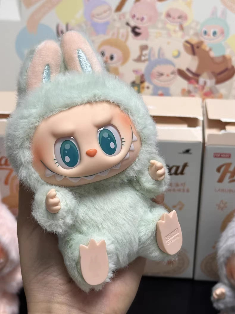 A hand holds a POP MART Labubu “Ququ” Plush Keychain – Have a Seat Series Green Bunny by The Monsters, showing its big-eyed doll face in a green bunny suit with ears, set among toy boxes and a cartoon-themed backdrop.