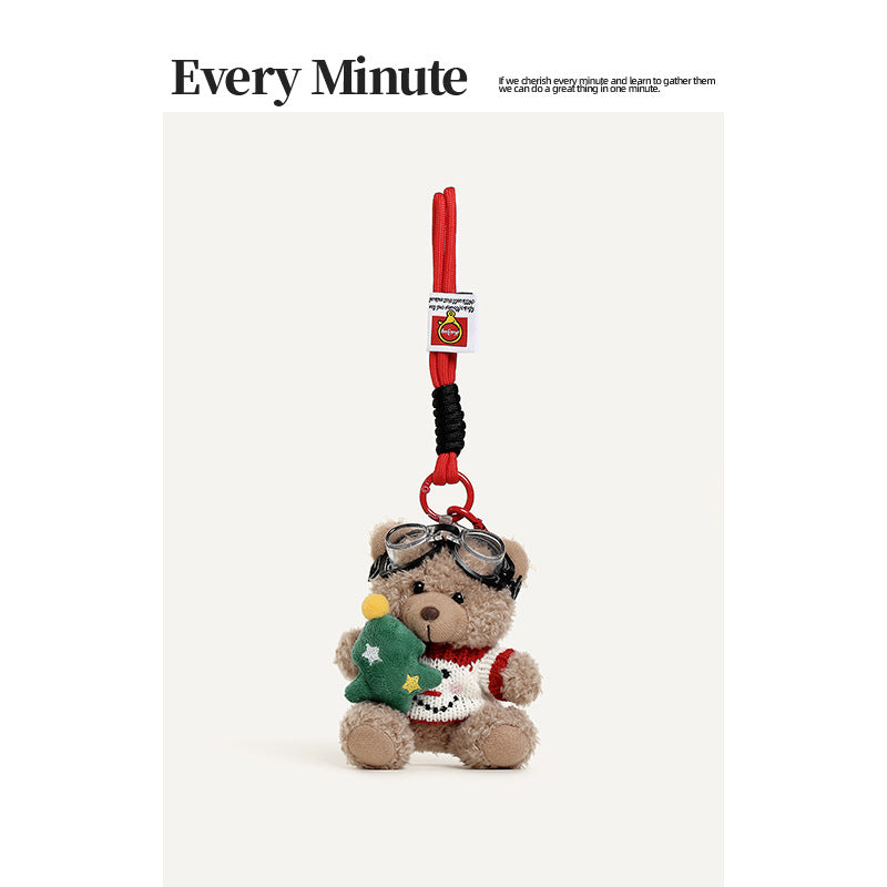 Beige plush teddy bear hanging charm in a white snowman-motif sweater holding a small green Christmas tree, with red strap an