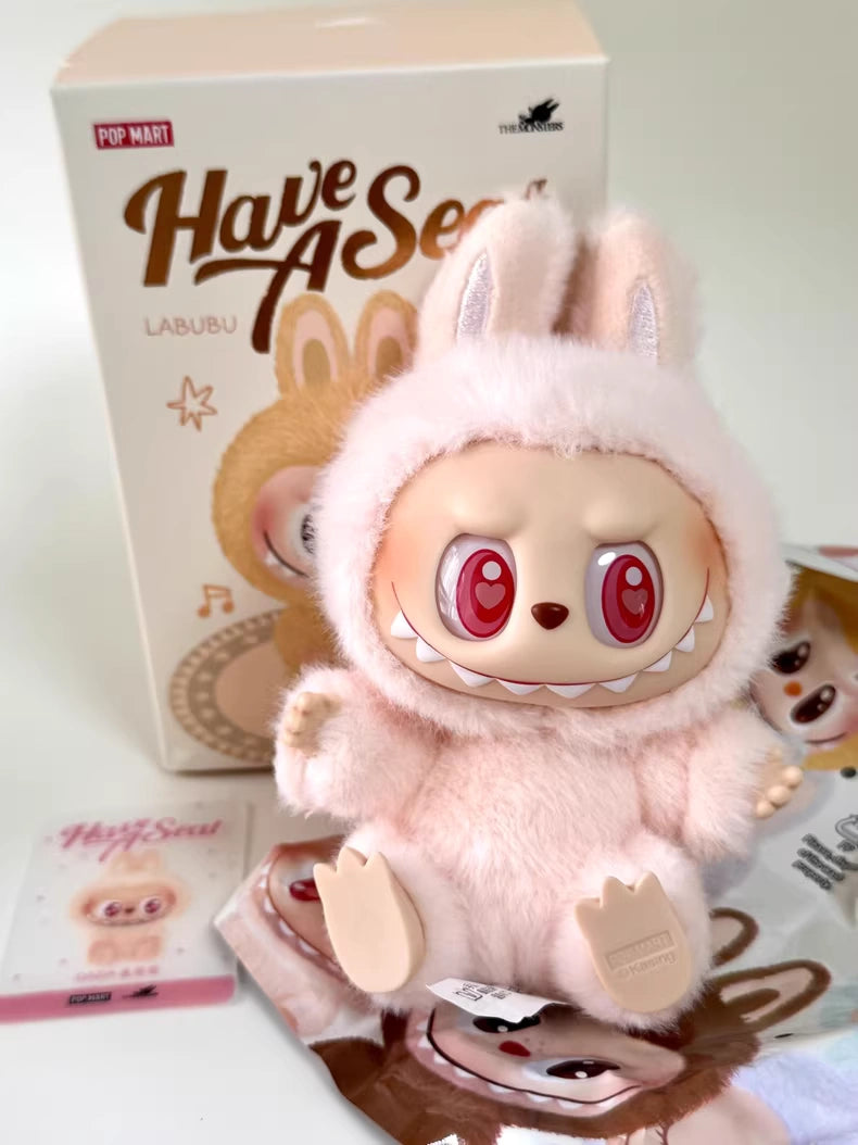 The Labubu “Dada” Plush Keychain – Have a Seat Series Pink Heart-Eye Bunny by POP MART The Monsters sits with its box and cards—a cute collectible perfect for fans.