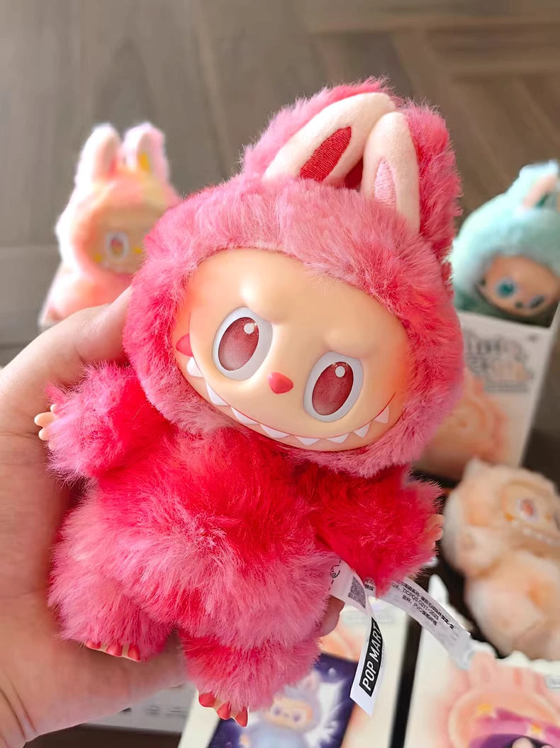 A hand holds a POP MART Labubu “Love” Plush Keychain – The Monsters “Big Into Energy” Series Red Bunny Bag Charm; other similar collectible toys in pink and blue outfits are visible in the background.