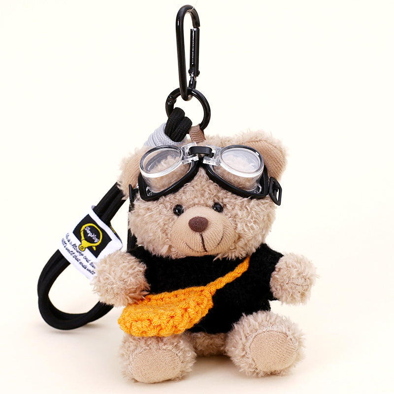 Beige teddy bear plush keychain in a black knit outfit with a small orange crossbody bag and aviator goggles