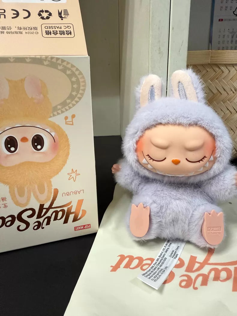 The Labubu “Zizi” Plush Keychain from the Have a Seat Series by POP MART’s The Monsters Collection sits beside its box and a printed fabric bag on a black surface.