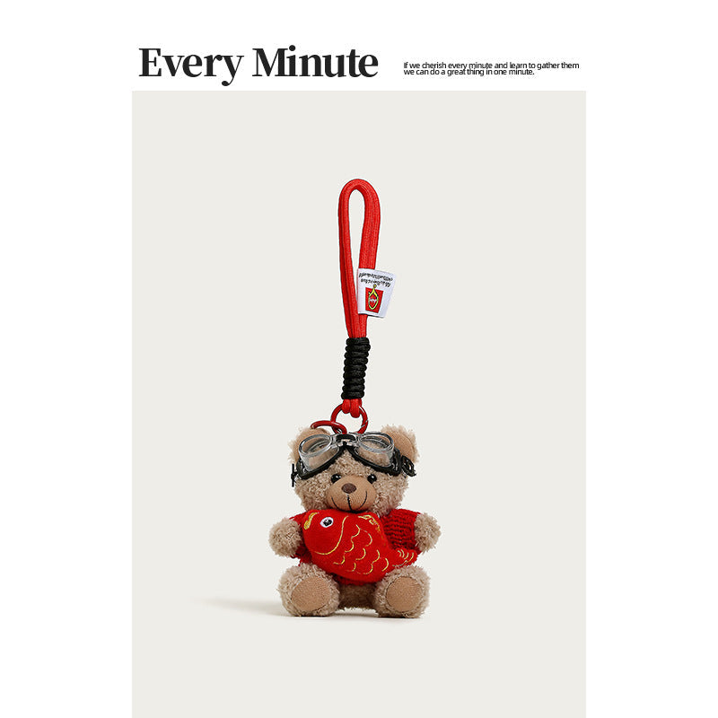 Brown plush teddy bear hanging charm in red sweater with goggles, holding a red koi fish plush, attached to red wrist-loop co