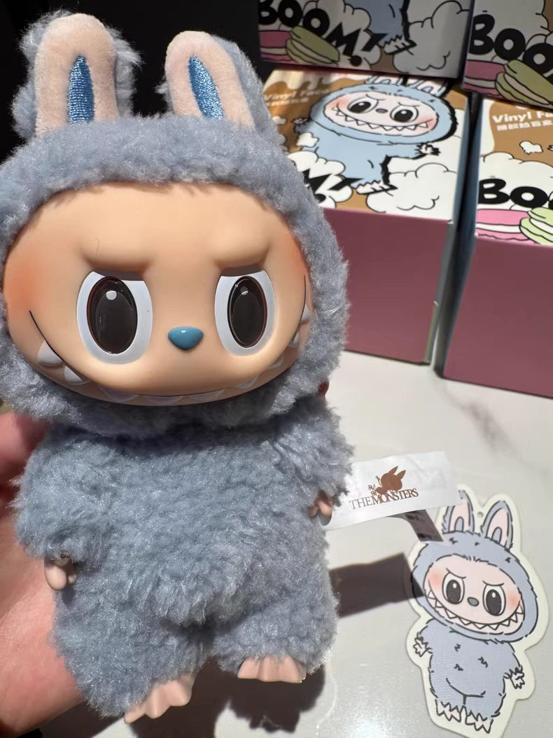 A hand holds a POP MART Labubu “Sea Salt Coconut” Plush Keychain—Standing Bunny in Blue Sherpa with big eyes and an angry expression; similar kawaii collectibles and illustrated boxes are seen in the background.