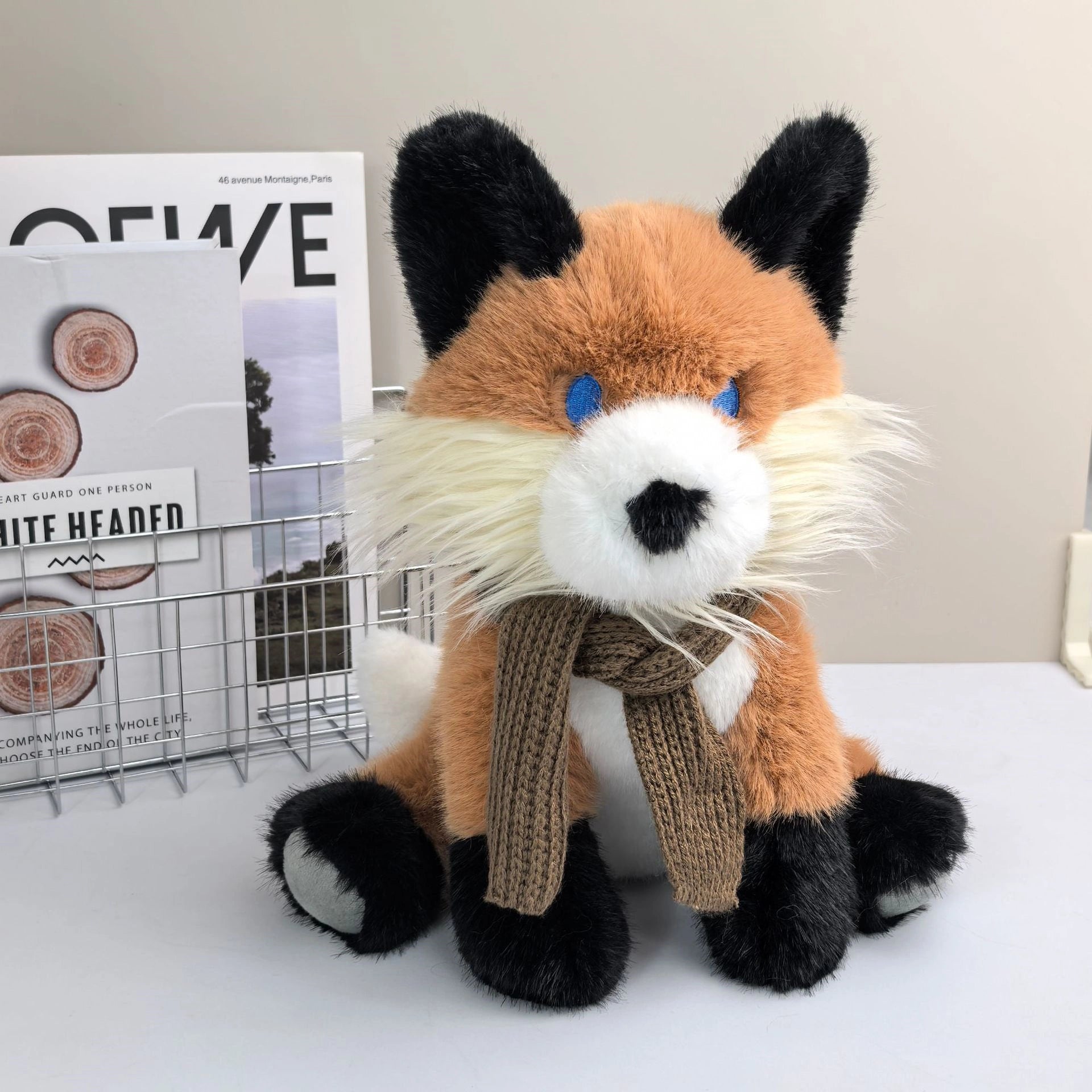 The ZoDoll Fox Plush Toy, a 30cm soft red fox stuffed animal with a brown scarf and white and black accents, sits on a desk near a metal magazine rack—ideal as charming woodland nursery decor.