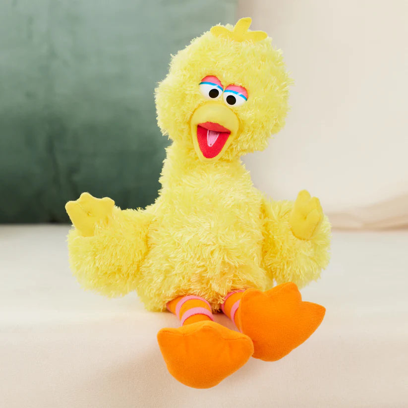 The ZoDoll Sesame Street Big Bird Plush – a 14-inch stuffed animal for kids and collectors – sits upright on a light surface, its bright yellow feathers, orange feet, and beak contrasting with a blurred green and beige backdrop.