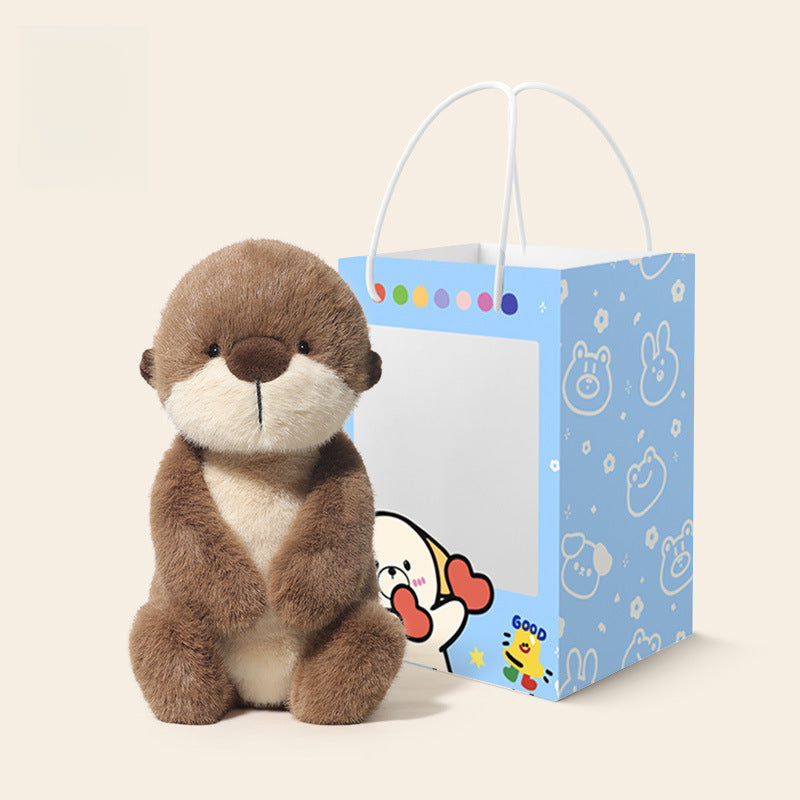 The JOOKI 36cm Otter Plush Toy, featuring soft brown and cream fur, sits by a blue gift bag with cartoon animals and hearts, creating charming woodland nursery decor on a light background.
