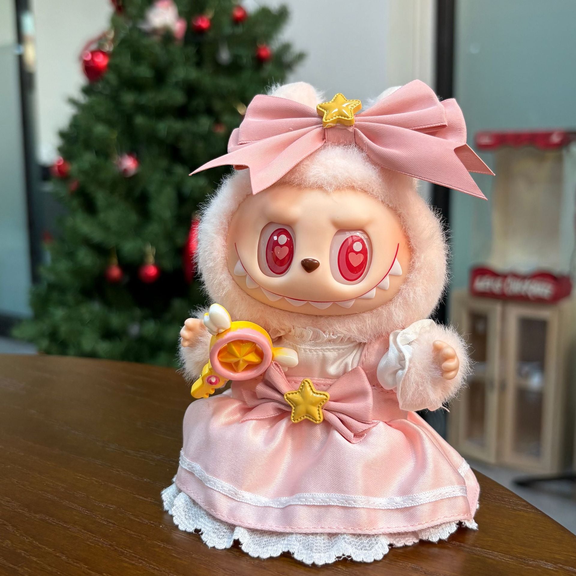 The ZoDoll Labubu Pink Magical Girl Satin Lace Dress with Star Wand and Bow Set features a doll with large red eyes and sharp teeth, dressed in pink with a hood, seated indoors by a decorated Christmas tree.