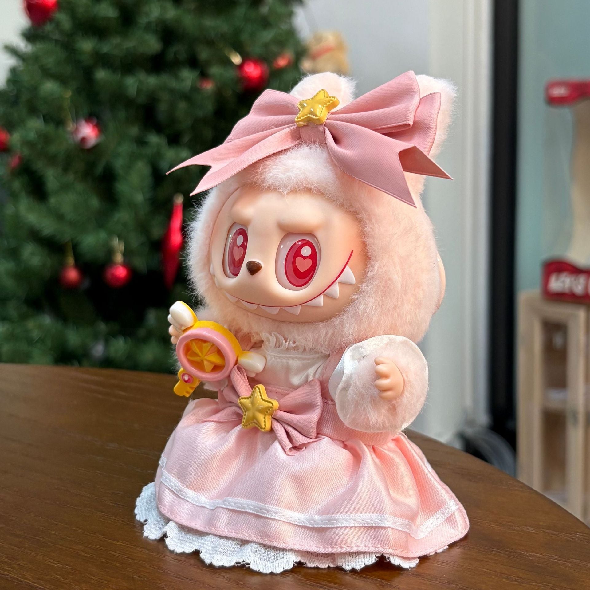 The ZoDoll Labubu Pink Magical Girl Satin Lace Dress with Star Wand and Bow Set doll, featuring wide red eyes, sits on a wooden surface with a decorated Christmas tree in the background.
