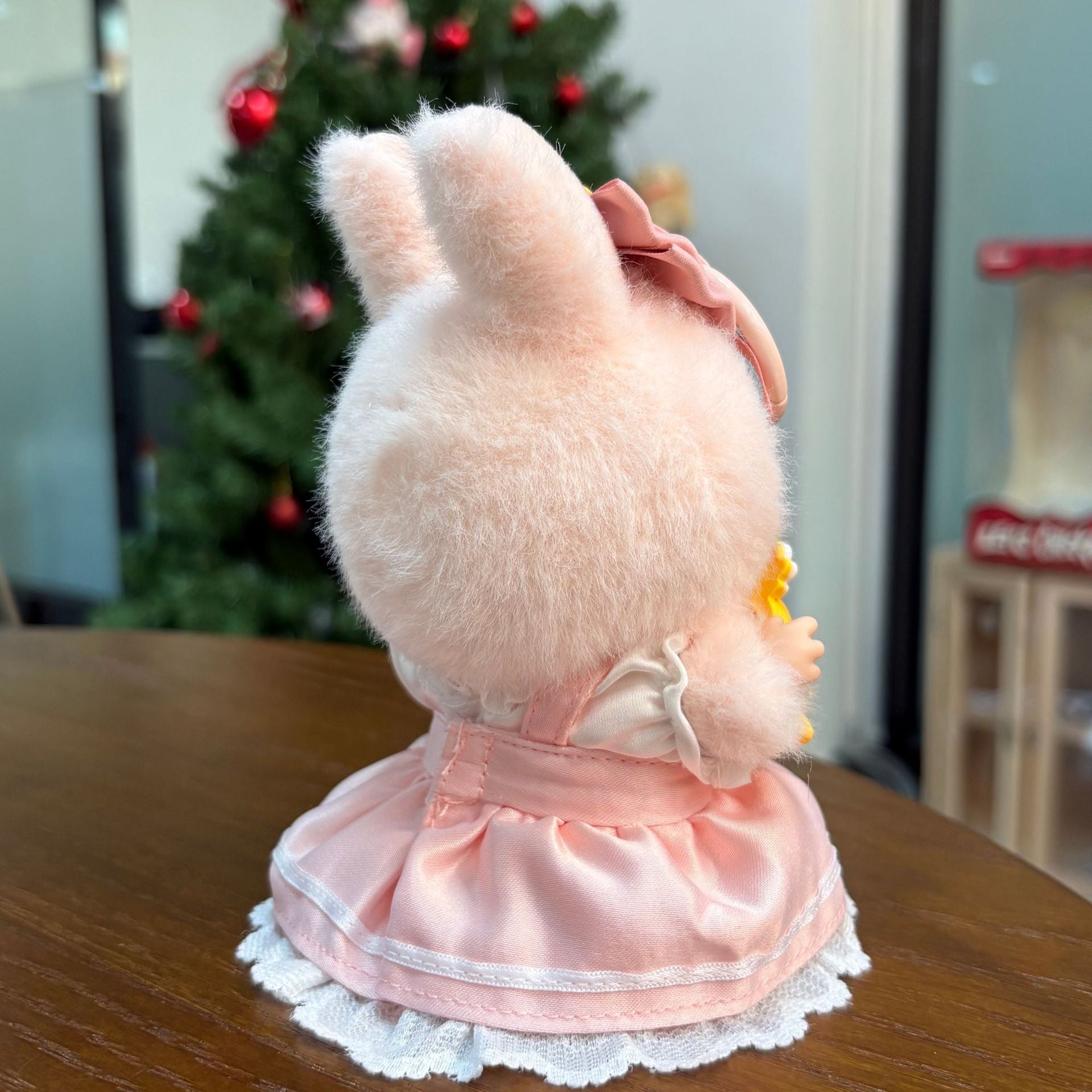 A ZoDoll Labubu plush wears a Pink Magical Girl Satin Lace Dress with Star Wand and Bow Set, sitting on a table with its back to the camera. A twinkling Christmas tree is visible in the background.