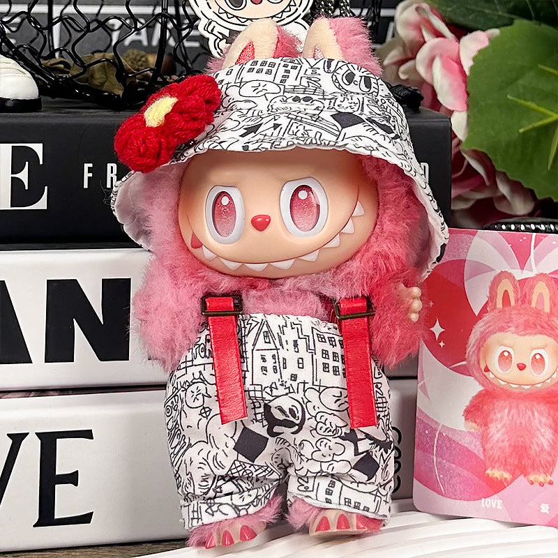 A ZoDoll Labubu plush wears the Graffiti Print Overalls Set with hooded cap and red suspenders, standing before books and a cup featuring matching character art, inspired by popular Labubu doll clothes.