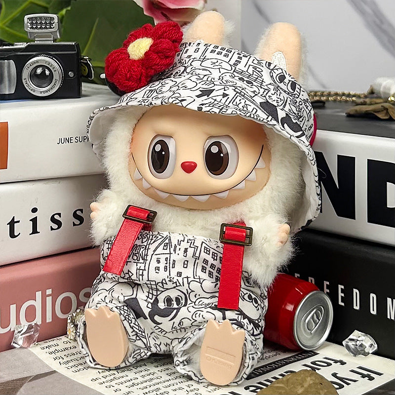 The ZoDoll Labubu Graffiti Print Overalls Set with Hooded Cap and Red Suspenders features a plush toy with large eyes, wearing a flower hat, sitting on books beside a small camera and an open soda can.