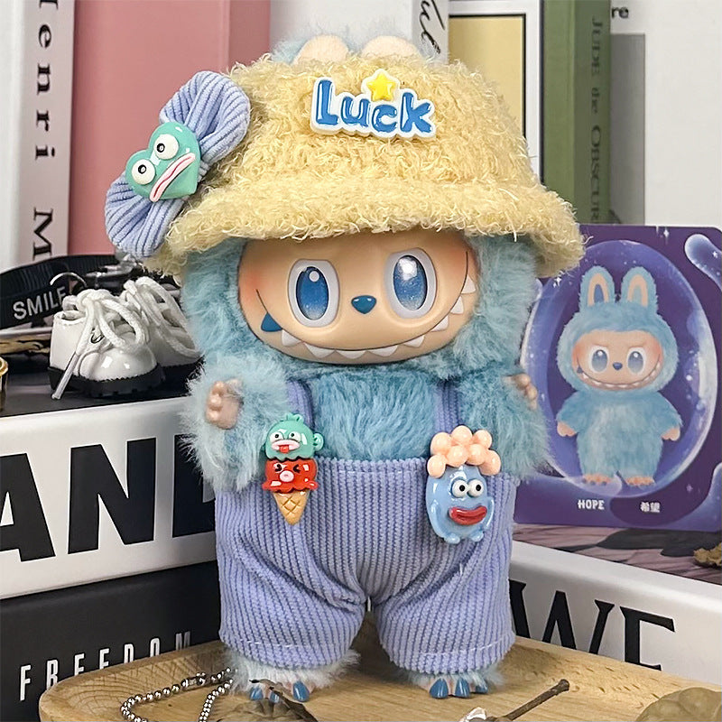 The ZoDoll Labubu Doll Lucky Bucket Hat and Corduroy Overalls Outfit Set features a plush blue and beige figure with big blue eyes, a yellow "Luck" hat, corduroy overalls, and cute pins—perfect among books and accessories.