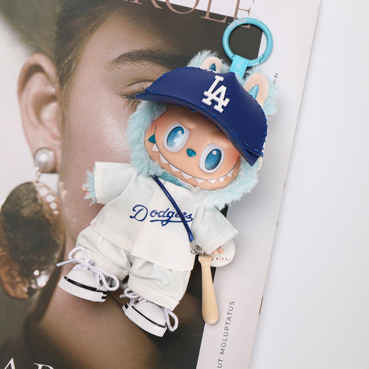 A ZoDoll Labubu Doll wearing a Dodgers baseball jersey and LA visor cap outfit set lies on a magazine, with a blue keychain attached to its head.