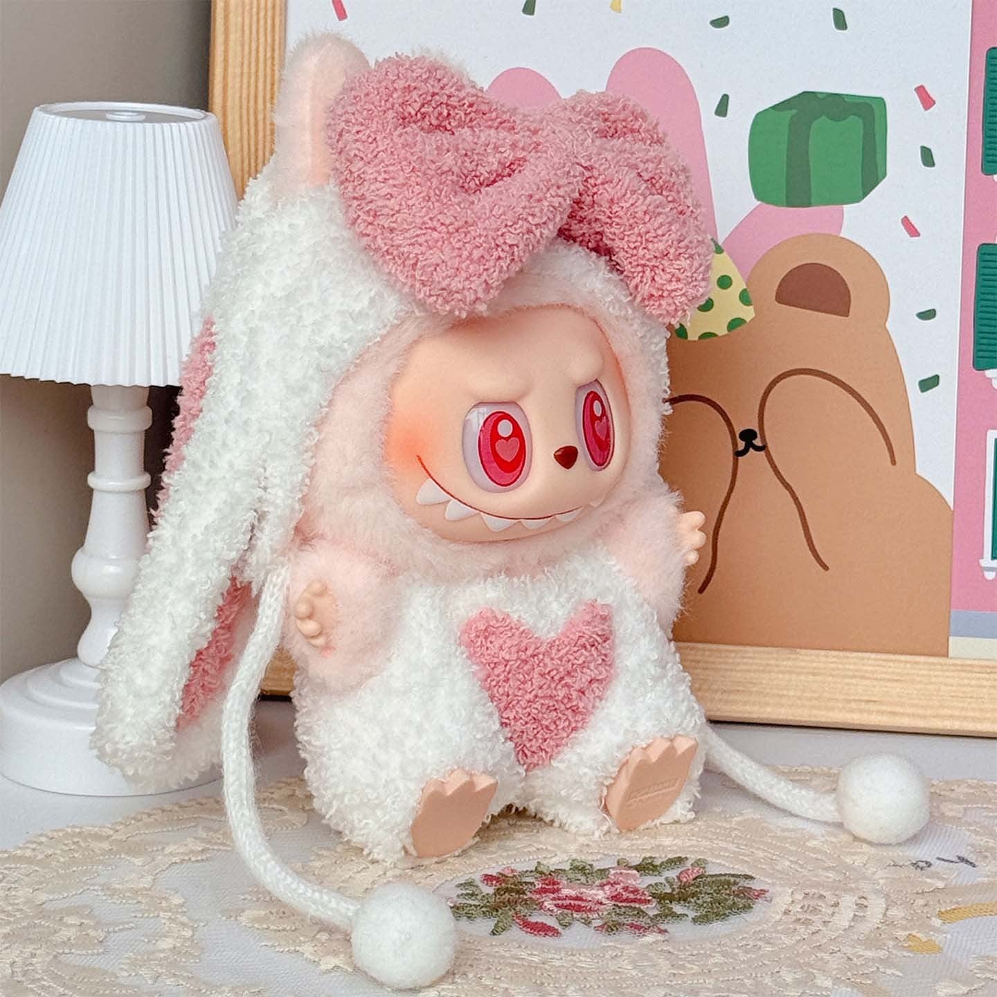 A ZoDoll Labubu Pink White Fluffy Bunny Onesie Plush Doll sits on a lace-covered table near a small lamp and framed art, featuring red eyes, pink cheeks, a heart on its chest, and a toothy grin.