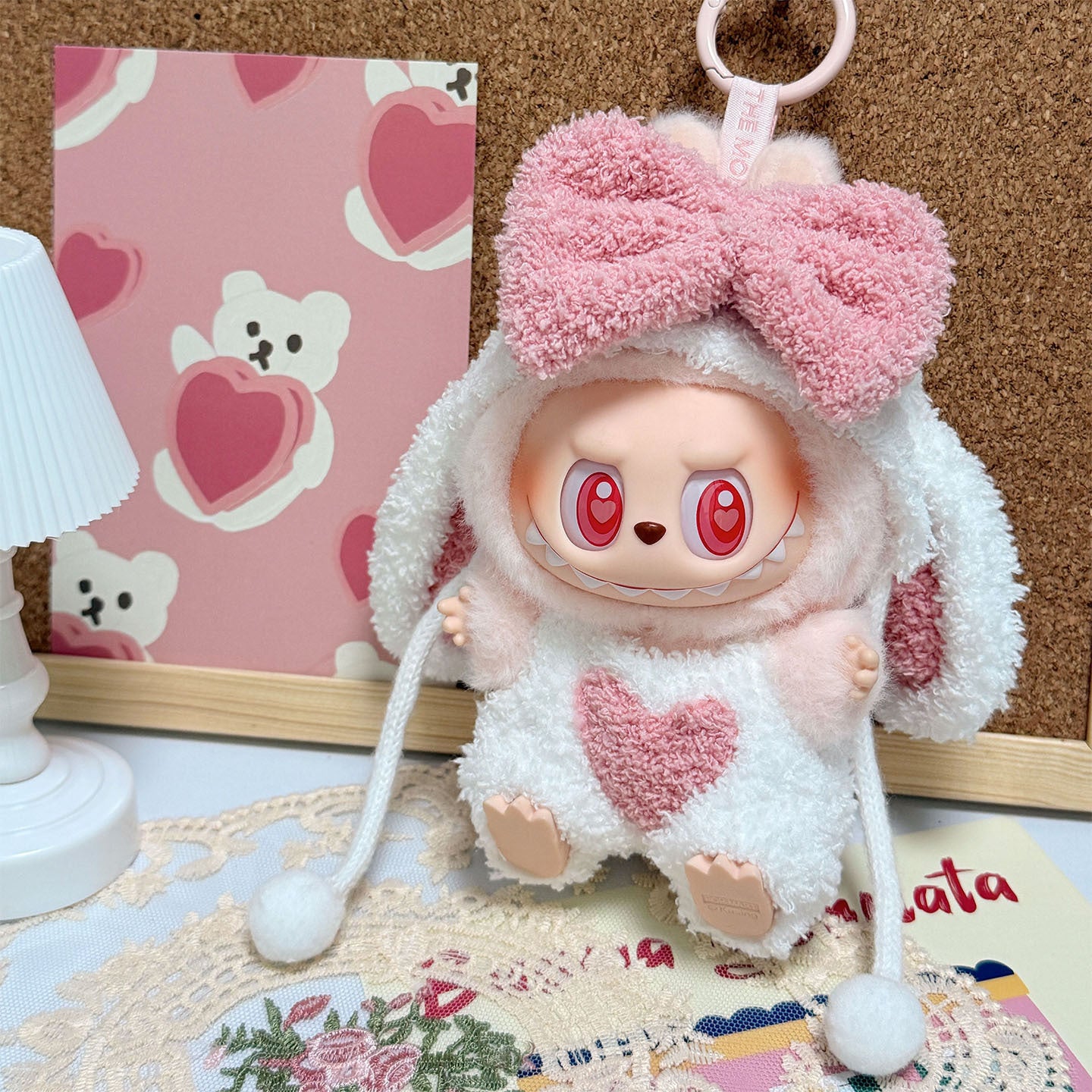 A ZoDoll Labubu Pink White Fluffy Bunny Onesie Plush Doll Costume, featuring a large pink bow and heart accents, sits on a decorated table beside a small lamp and a teddy bear-themed postcard.
