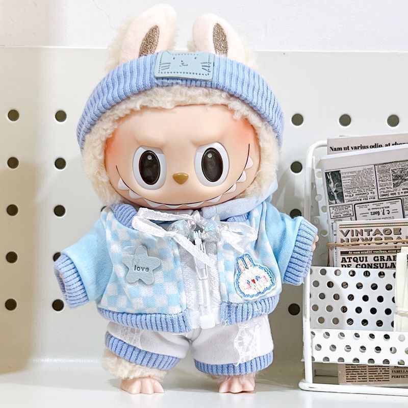 A ZoDoll Labubu doll wears the Blue Checkered Jacket and Knit Beanie Outfit Set, standing beside a metal rack holding newspapers.