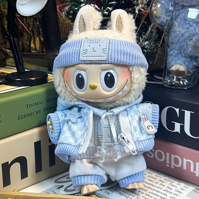A ZoDoll plush toy with big eyes and a wide grin wears the Labubu Blue Checkered Jacket and Knit Beanie Doll Outfit Set, standing on stacked books next to a transparent bear figurine.