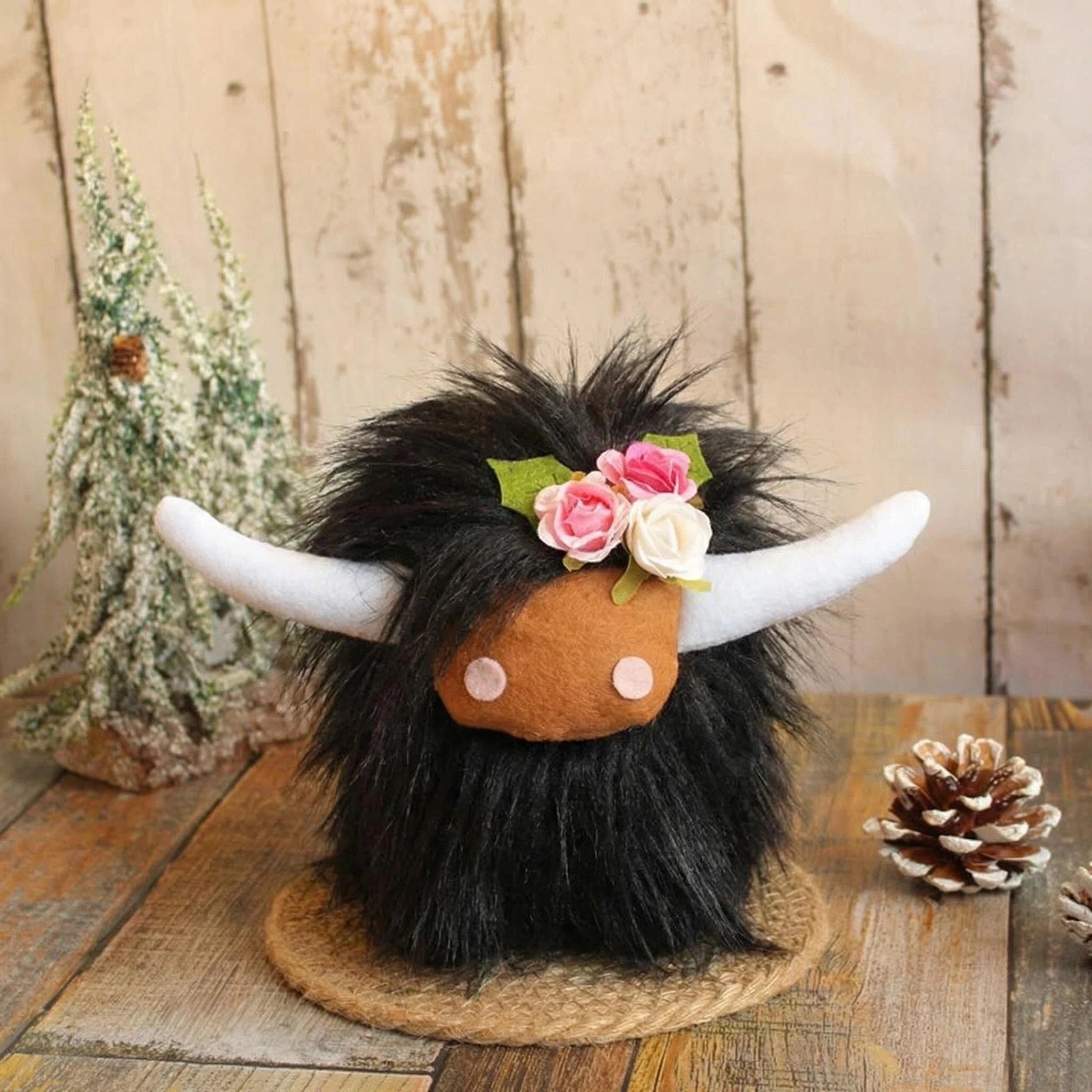 The ZoDoll Highland Cow Plush Toy with Pink Bow, featuring soft long black fur and a flower crown, sits on a mat beside a pinecone and small artificial tree on a wooden surface.