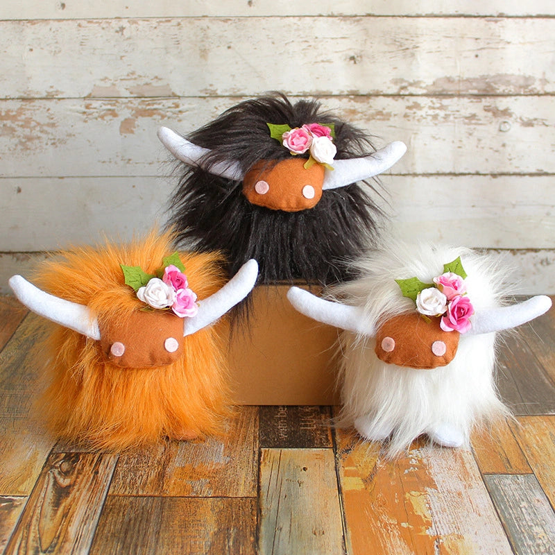 Three ZoDoll Highland Cow Plush Toys with pink bows, in black, orange, and white, are displayed on and in front of a small box against a rustic wooden background. These cuddly plush cows feature soft long hair and come in six colors.
