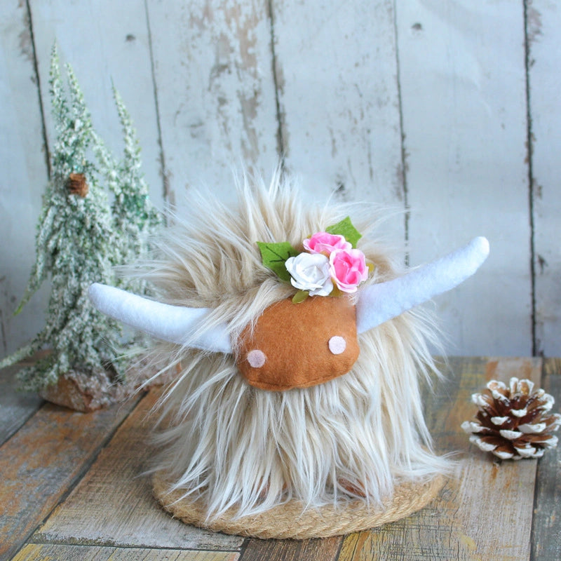 A ZoDoll Highland Cow Plush Toy with a pink bow and soft long hair sits on a wooden surface beside a pinecone and a small frosted tree. Available in 6 colors.