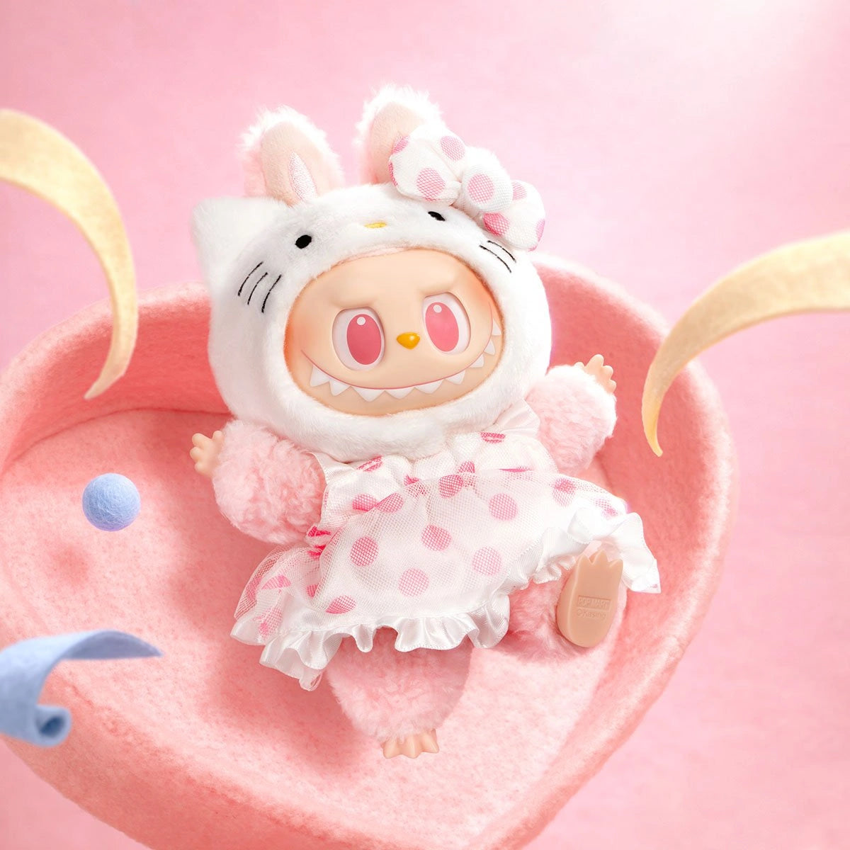 A doll in a pink and white bunny costume with a polka dot dress lies on a pink surface with curved yellow shapes, a small blue ball, and POP MART’s LABUBU Hello Kitty and Friends Series Hello Kitty Plush Pendant Figure nearby.