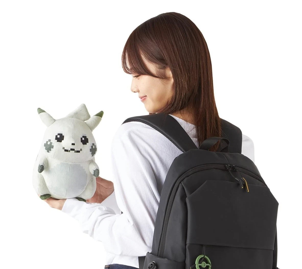 A woman with a black backpack holds the Retro 8-Bit Pikachu of Beginnings Plush by ZoDoll, a Classic Chunky Pokémon 30th Anniversary toy, looking at it while standing against a plain white background.