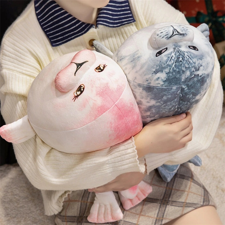 A person in a cream sweater holds two ZoDoll Seal Plush Toys—chubby seal pillows in pink/white and gray/white—while sitting on a carpeted floor. Available as soft stuffed animal pillows, 20cm or 40cm.