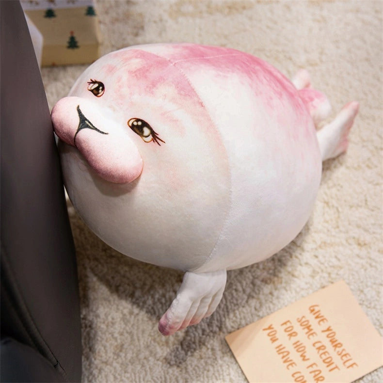 A ZoDoll Seal Plush Toys Cute Chubby Seal Plush Pillow, pink and white with expressive eyes, sits on a black surface atop a beige carpet near a card with motivational text. Size options: 20cm or 40cm.