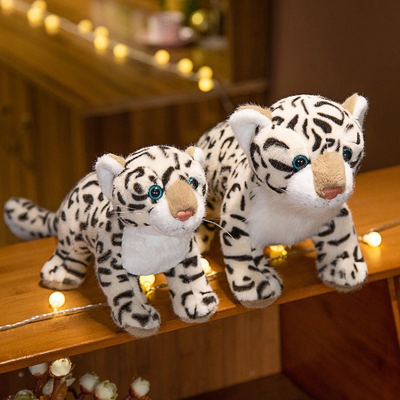 Two ZoDoll Snow Leopard Plush Realistic Stuffed Toys (43cm & 52cm), featuring black spots and blue eyes, rest on a wooden surface softly lit by string lights in the background.