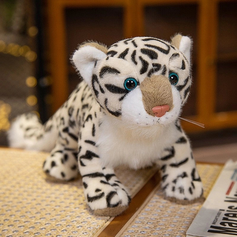 A ZoDoll Snow Leopard Plush Realistic Stuffed Toy (43cm/52cm) with black stripes, blue eyes, and a pink nose sits on a wooden surface beside magazines.