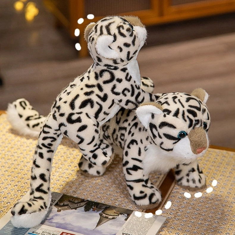 Two ZoDoll Snow Leopard Plush toys (43cm, 52cm), with realistic black spots, are stacked playfully on a table near a magazine, creating a lively scene with these soft wild cat stuffed animals.