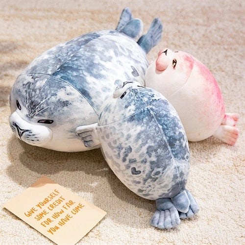 Three ZoDoll Seal Plush Toys, soft and chubby at 20cm and 40cm sizes, rest on a carpeted floor next to a note that says, "Give yourself some credit for how far you have come.