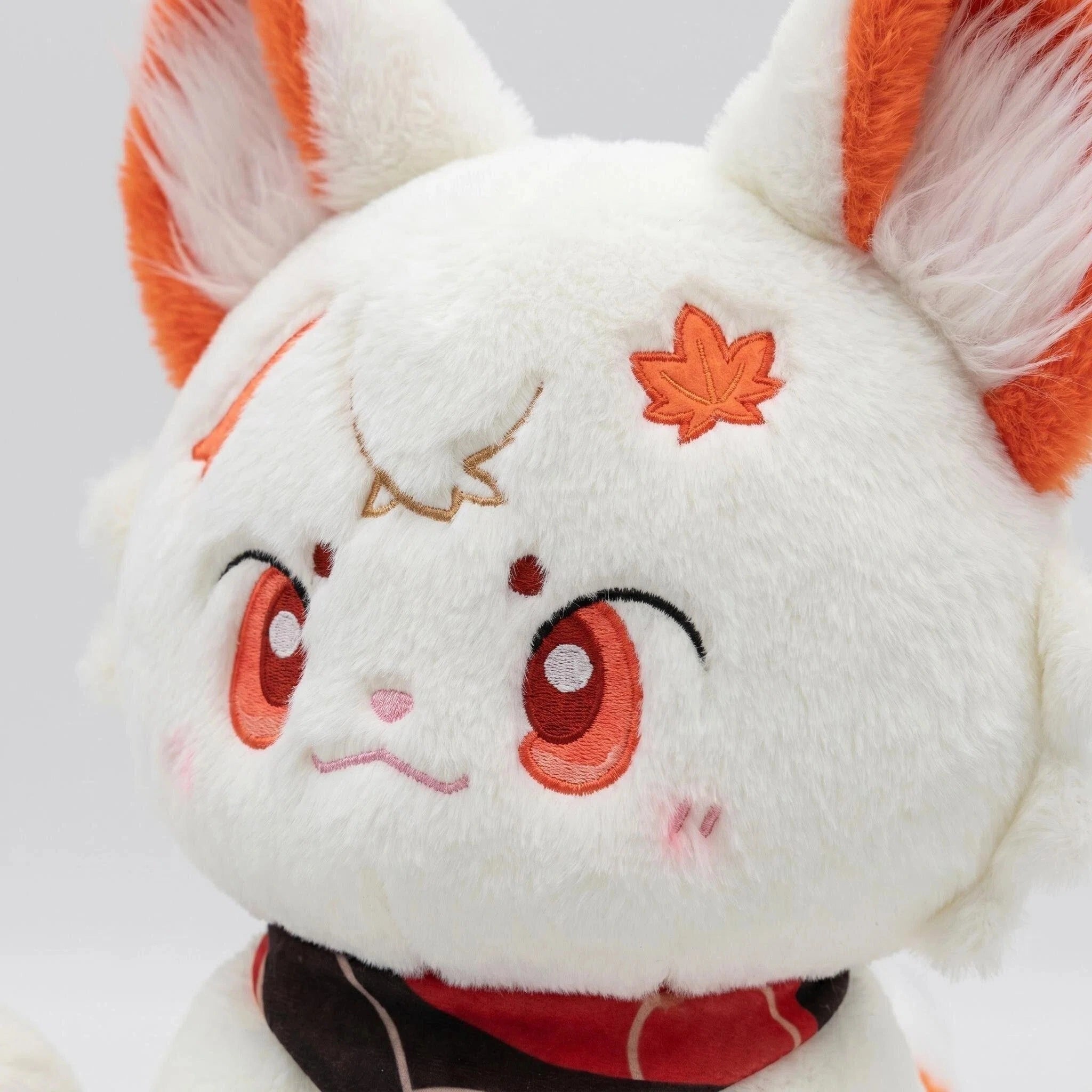 The ZoDoll White Fox Plush Toy is a cute plush doll with orange ears, a kawaii embroidered face, a maple leaf on its head, and a red bandana around its neck.
